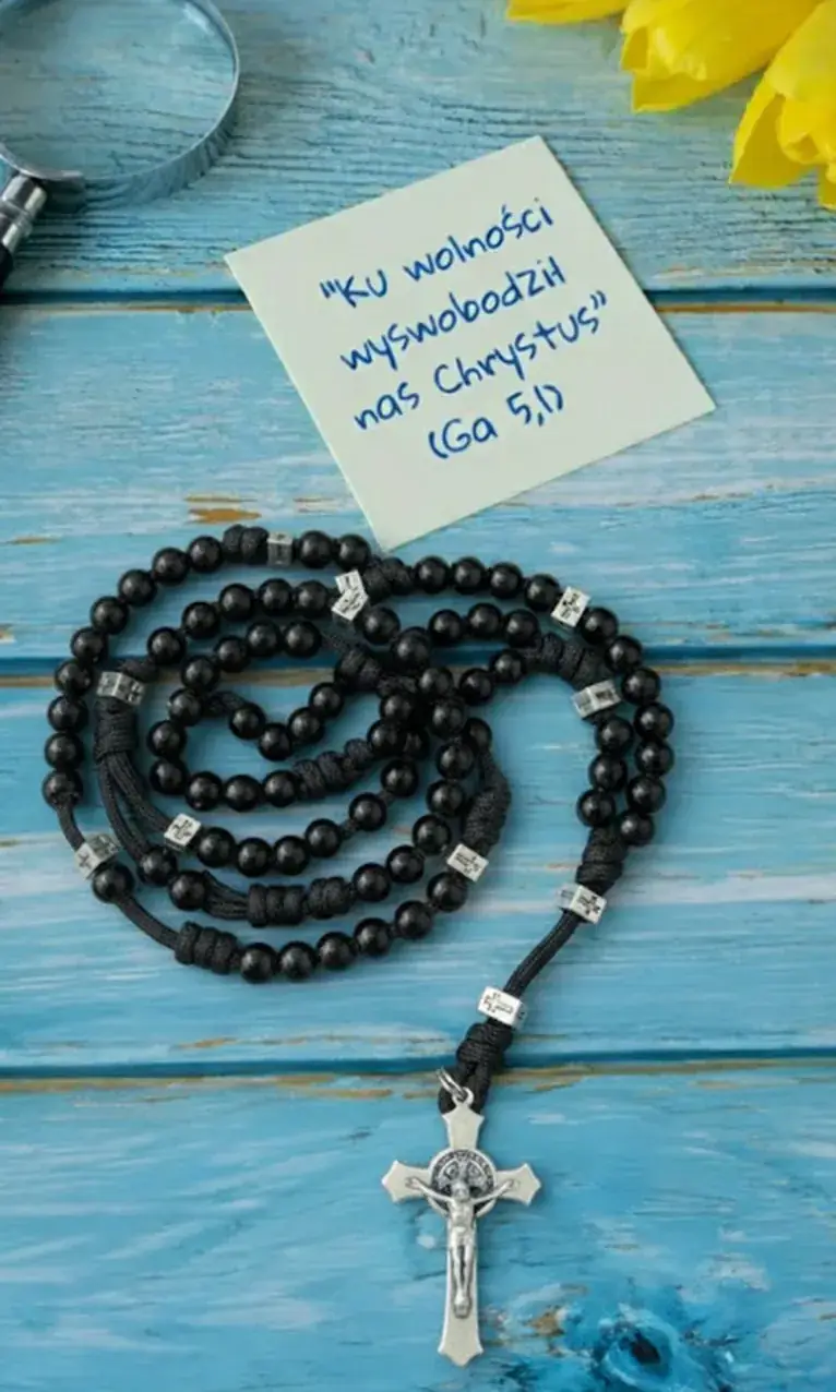 Różaniec z krucyfiksem na niebieskim drewnianym stole i kartka z cytatem: Ku wolności wyswobodził nas Chrystus (Ga 5,1)